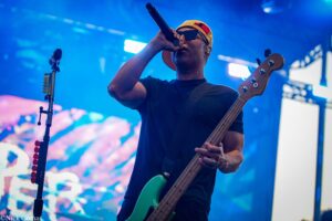 Male guitarist on stage wearing a yellow cap and sunglasses, singing into a microphone while playing a green bass under blue lights.
