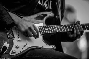 Close-up of a guitarist's hands on a Strat-style electric guitar, leather jacket visible in the frame.
