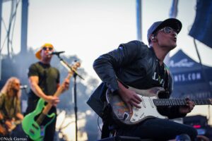 Guitarist in a black leather jacket, cap, and sunglasses plays a white electric guitar on stage with band members blurred in the background.