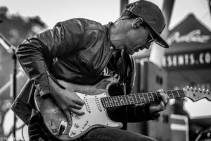 Black-and-white photo of a guitarist wearing a cap and leather jacket, playing a white Fender Stratocaster on stage.
