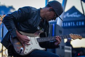 Guitarist in a black leather jacket and cap, playing a white Fender Stratocaster on stage.