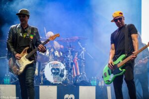 Two musicians perform on a smoky blue stage: left guitarist in a cap and leather jacket, right bassist in a yellow cap and black shirt with a green bass; drum set visible in back with 'Pepper' on the bass drum.