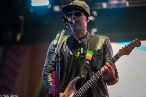 Guitarist wearing a leather jacket and cap sings into a microphone during a live performance, with a colorful guitar strap visible.