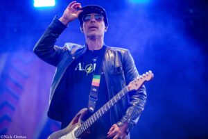 Guitarist on stage wearing a black leather jacket, cap and sunglasses, adjusting a hat while playing an electric guitar under blue stage lights.