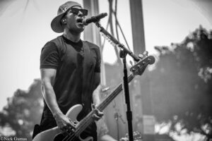 Bassist wearing a cap and sunglasses sings into a microphone while playing a bass on stage (black-and-white photo).