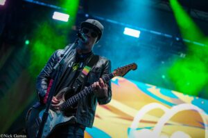 Guitarist performing on stage in a black leather jacket, cap, and sunglasses under bright neon lights.