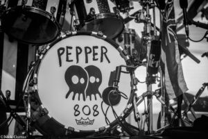 Close-up of a band's drum kit bass drum with 'PEPPER' and a skull graphic is pictured in a black-and-white shot.