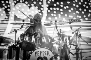 Black-and-white shot of a bearded drummer wearing a cap, mid-performance behind a large drum kit with 'PEPPER' on the bass drum.