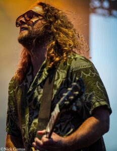 Guitarist with long curly hair and beard wearing sunglasses, playing a guitar on stage in a patterned shirt.