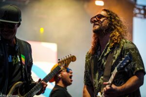 Two guitarists on stage, one with dark sunglasses and long hair, the other wearing a cap and leather jacket, playing electric guitars under warm lighting.