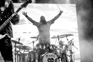 Bearded drummer with tattoos raises both arms above a drum kit on stage, bass drum shows 'PEPPER' logo