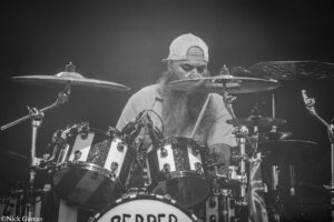 Black-and-white photo of a bearded drummer wearing a backward baseball cap behind a large drum kit on stage.