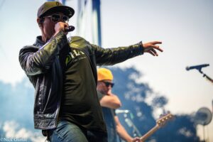 Male singer in a black leather jacket and cap performs on an outdoor stage, microphone in hand with a guitarist in the background.