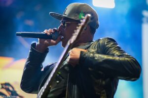 Male singer wearing a cap and sunglasses performs on stage, holding a microphone and guitar in hand on a live show