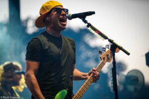 Male bassist singing into a microphone on stage, wearing a yellow backwards cap and dark sunglasses, with a green bass guitar in view.