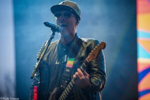 Male guitarist singing into a microphone on stage, wearing sunglasses and a brown cap with a leather jacket, playing an electric guitar amid stage lighting and fog.
