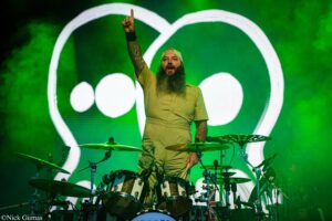 Drummer with a long beard raises one finger while standing behind a full drum kit on a green-lit stage.