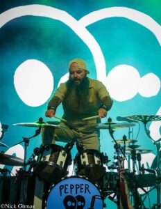Drummer with a long beard and backward-cap playing a drum kit on a brightly lit stage, with a large blue backdrop behind him and the word 'PEPPER' on the bass drum.