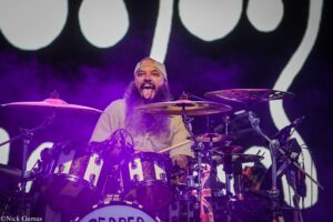 Drummer with long beard and a backward cap playing a large drum kit on stage bathed in purple light.