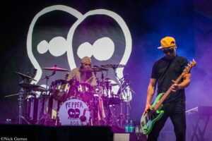 Drummer with a beige cap playing a drum kit on stage, guitarist in a yellow cap on the right; a large white logo on a dark screen behind them.