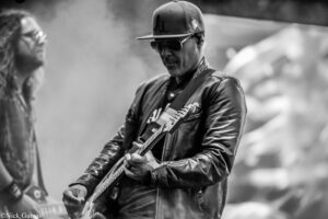 Black-and-white photo of a guitarist wearing a cap and sunglasses, playing an electric guitar on stage.