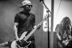 Male bassist wearing a backwards cap and sunglasses plays a white bass on stage, other guitarist visible in background.