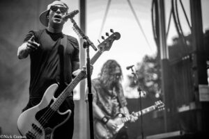 Black-and-white photo of a bass guitarist singing into a microphone on stage, with another guitarist in the background.