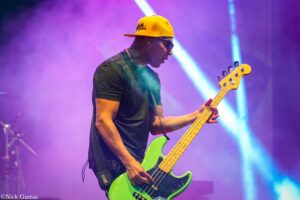 Musician wearing a yellow cap and sunglasses plays a bright green bass guitar on a purple-lit stage for a live performance.
