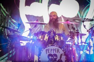 Drummer with a long beard and yellow cap performs on stage behind a drum kit with a skull-themed bass drum reading 'PEPPER' on the front.