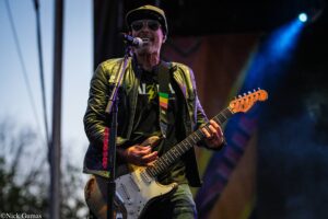Male guitarist on stage wearing a yellow cap and sunglasses, singing into a microphone while playing a white electric guitar.
