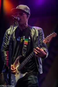 Male guitarist performing on stage, wearing a beige cap, sunglasses and a black leather jacket, singing into a microphone.