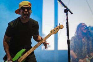 Bassist wearing a yellow cap and dark sunglasses plays a neon-green bass guitar on a blue-lit stage, with a microphone stand nearby.