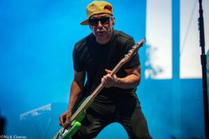 Guitarist wearing a yellow cap and sunglasses and playing a green bass guitar on a blue-lit stage.