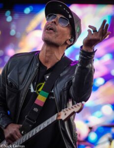 Guitarist wearing a black cap and sunglasses performs on a stage with bright, colorful lights in the background.