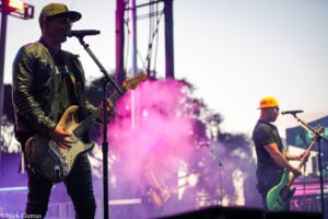 Guitarist in a green leather jacket and cap sings into a microphone on stage while playing a white electric guitar; pink stage smoke around him.
