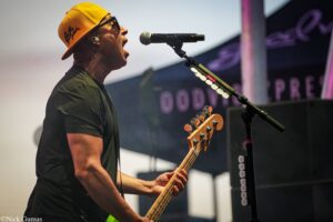 Male bassist wearing a yellow cap sings into a microphone on stage while playing a bass guitar.