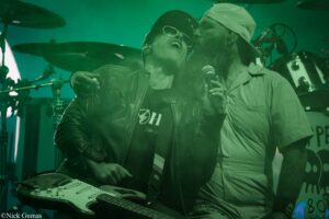 Two musicians on a stage bathed in green lighting: a leather-jacketed guitarist playing an electric guitar while a bearded singer with a microphone leans in.