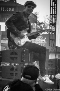 Male guitarist wearing a cap and sunglasses, playing a white Fender Stratocaster on a stage during a live performance.