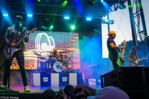 Band on an outdoor stage performing live with colorful lights, guitarists left and right and a drummer in the center; audience visible in foreground.