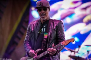 Guitarist performing on a vibrant stage, wearing sunglasses, a maroon leather jacket, and a cap with 'hi' on it while playing an electric guitar. (Live concert)