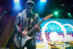 Guitarist in leather jacket and sunglasses performs on a lit stage, singing into a microphone with a worn white electric guitar.