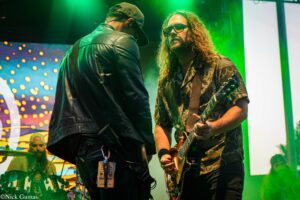 Guitarist with long hair and sunglasses playing a guitar on a green-lit stage, another musician facing away in a leather jacket nearby.