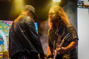 Two musicians on a dim stage lean toward each other; one wears a black leather jacket, the other has long hair, sunglasses, and is holding a guitar.