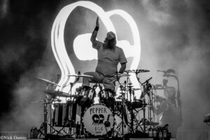 Drummer on stage raising one hand while playing a complex drum kit, with a large circular Pepper logo backdrop behind him (black-and-white concert photo).