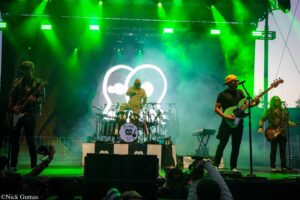 Live band performing on a neon-green illuminated stage; drummer center, guitarist left, bassist right with yellow cap.