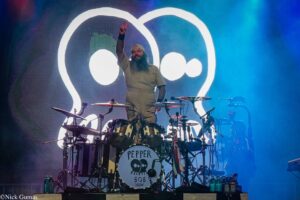 Drummer on stage raising a peace sign, large white logo backdrop, Pepper 808 bass drum, blue lighting nearby.