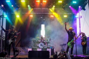 Band performs on an outdoor stage under bright multi‑color lights; guitarist left, lead singer with yellow cap, drummer center, audience in front.