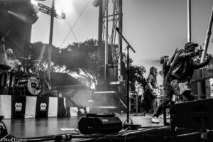 Live rock band on stage: drummer left, guitarist to the right, vocals at mic, black-and-white festival setup with skull banners and stage rigging