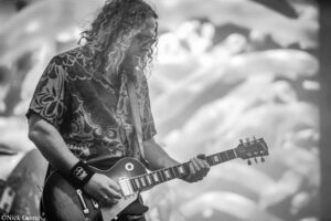 Guitarist with long curly hair and a patterned shirt playing a sunburst electric guitar on stage.