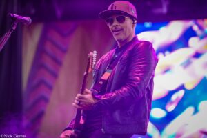 Guitarist wearing a cap, sunglasses, and a purple leather jacket playing an electric guitar on a purple-lit stage.